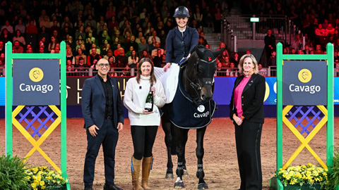 Equestrian rider on a horse with three people beside them in an indoor arena, with 'Cavago' branding and a seated audience in the background.